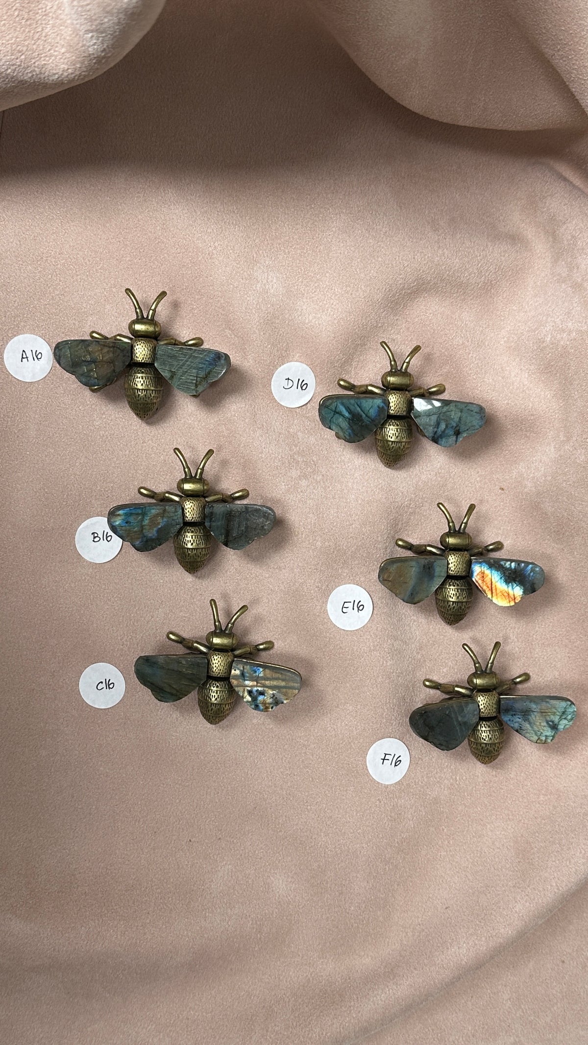 Six small bee-shaped labradorite pieces arranged on a soft beige fabric background, each with metallic gold bodies and blue-gold wings.